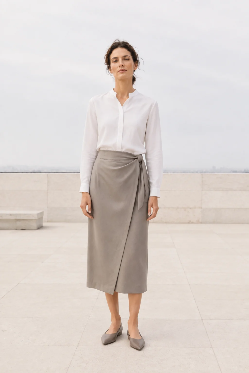 Woman in a clean white shirt and cool-taupe wrap midi skirt on a bright city terrace